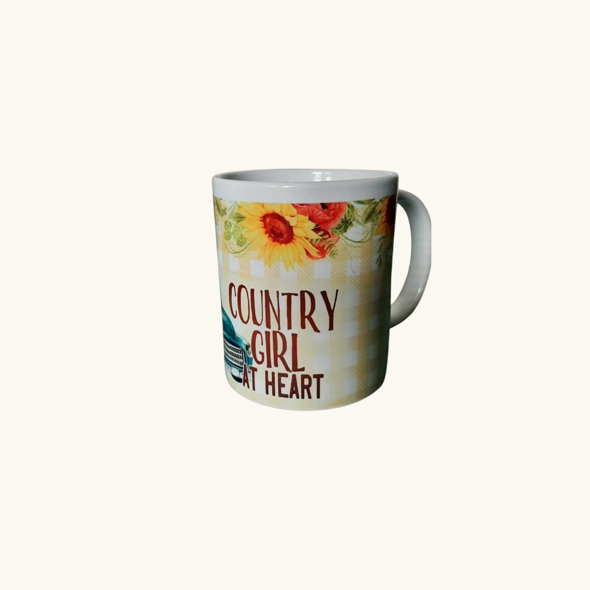 A white ceramic coffee mug with a rustic country-themed design featuring a sunflower, red flowers, a plaid pattern, and the text 'COUNTRY GIRL AT HEART' in brown lettering, set against a solid off-white background.