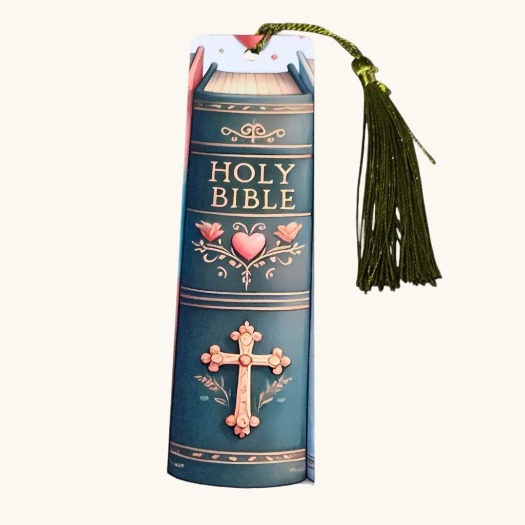 A Holy Bible book-shaped bookmark with a green tassel, featuring a dark teal cover design with gold lettering and pink heart and cross illustrations, shown on a light cream background.