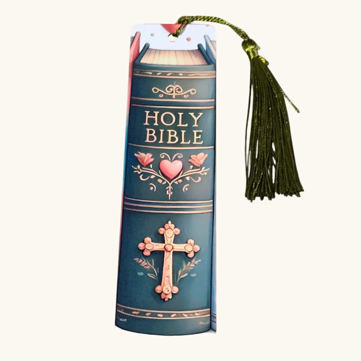 A Holy Bible book-shaped bookmark with a green tassel, featuring a dark teal cover design with gold lettering and pink heart and cross illustrations, shown on a light cream background.