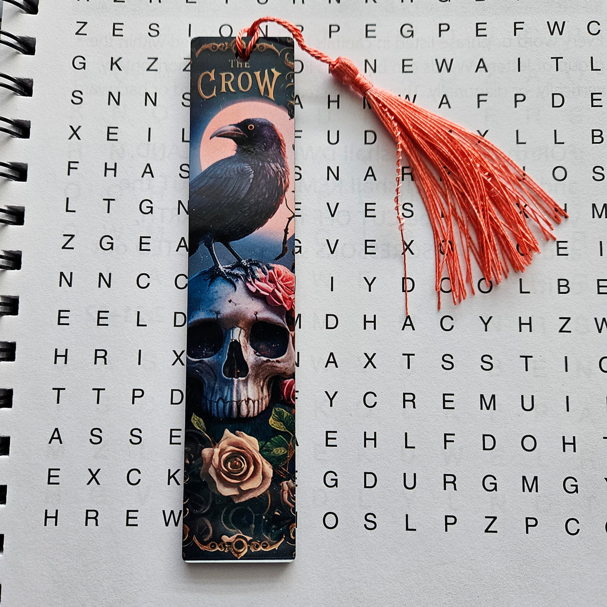 A gothic-themed bookmark featuring a raven perched on a skull with roses, complete with a red tassel, laid flat on a word search page.