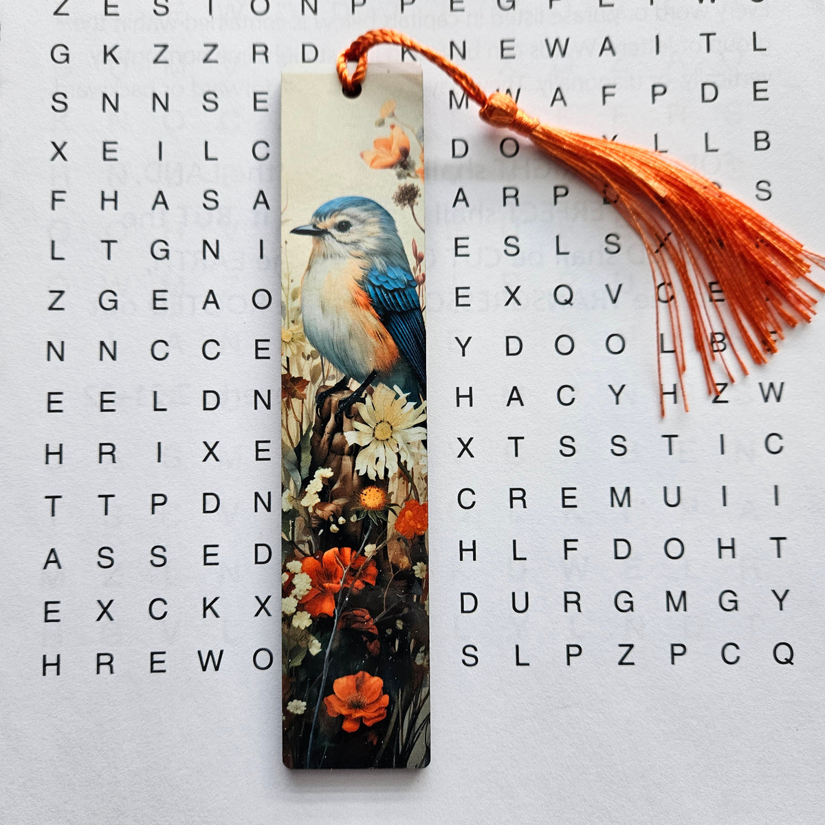A rectangular wooden bookmark with a vibrant painted bird perched among flowers, featuring a bright orange tassel, displayed on a white word-search sheet.