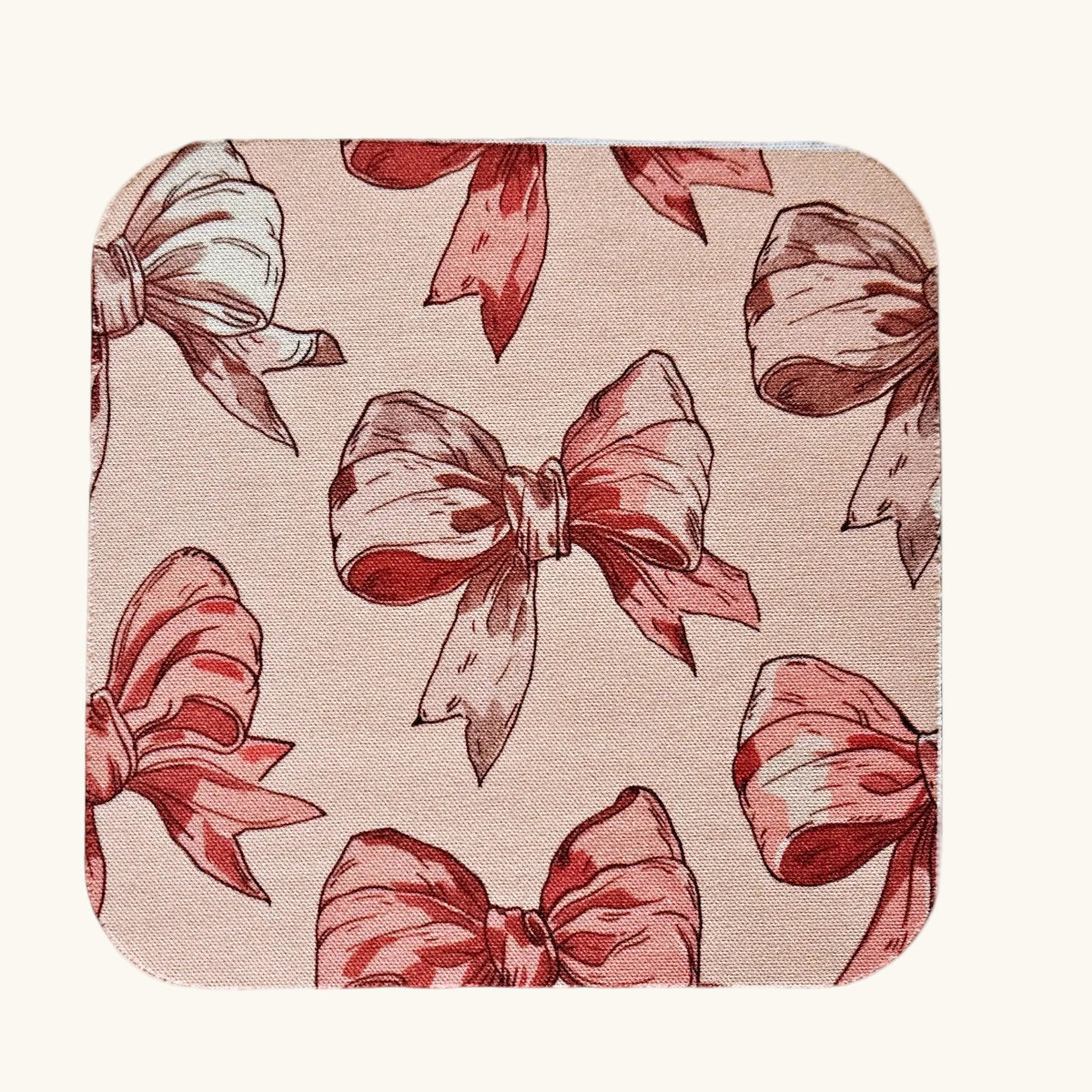A square fabric item with rounded corners, featuring a repeating pattern of red and dusty rose bows on a light beige textured background, shown against a solid cream backdrop.