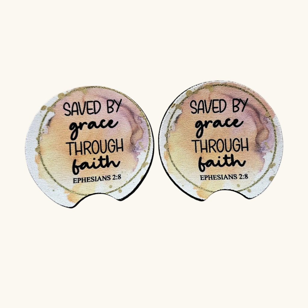 Two oval-shaped coasters with a watercolor design in soft peach, beige, and gold tones, featuring the text 'SAVED BY grace THROUGH faith EPHESIANS 2:8' in black lettering, shown on a light cream background.